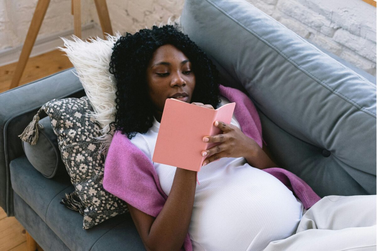 A pregnant woman reading a book on a couch, immersed in a pregnancy journal filled with prompts.