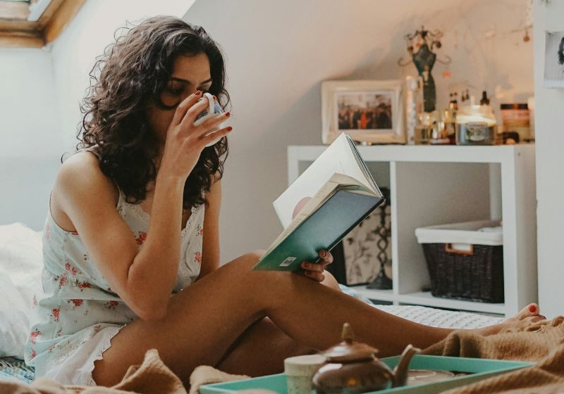 An image of a woman relaxing and reading while sipping a drink as a part of her self care routine